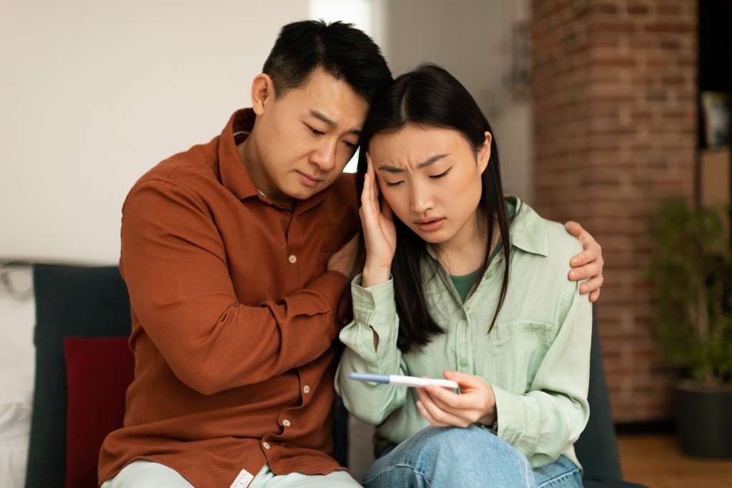 Upset mature asian husband comforting his depressed young wife with negative pregnancy test, sitting on sofa at home. Korean couple cannot have baby, suffering from infertility indoors